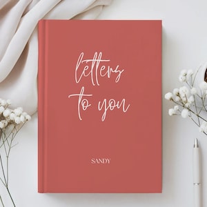 Op de afbeelding: Een rechthoekig notitieboek met harde kaft in een effen terracotta rode kleur. De voorzijde bevat de witte cursieve tekst "letters to you" in het midden van de bovenste helft, met de naam "SANDY" in witte hoofdletters onderaan.