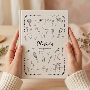 May include: A cream-colored hardcover recipe book featuring black line-art illustrations of kitchen utensils, cookware, and hearts. The text on the front cover reads Olivia's Recipe Book, enclosed within a decorative black border of swirling lines.