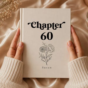 May include: A cream-coloured book with the words "Chapter 60" in black script. Below the text is a floral illustration and the name "Susan". The book is held by a person wearing a cream-coloured sweater.
