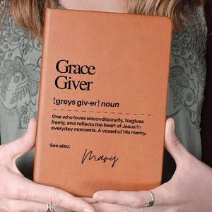 May include: A tan-coloured journal with the words "Grace Giver" and a definition printed on the cover. The definition reads, "One who loves unconditionally, forgives freely, and reflects the heart of Jesus in everyday moments. A vessel of His mercy." The name "Mary" is also printed on the cover.