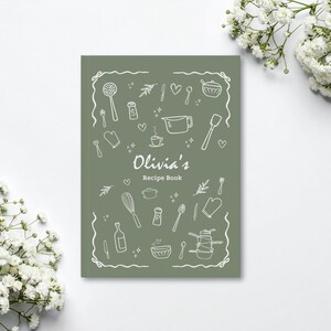 Puede incluir: Un libro de recetas verde salvia con el t&iacute;tulo "Olivia's Recipe Book" en una fuente manuscrita. La portada presenta dibujos lineales blancos de utensilios de cocina y alimentos, incluyendo un batidor, una cuchara y una taza medidora.