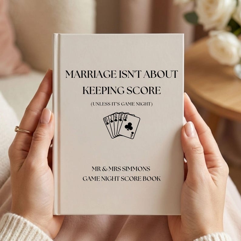 May include: A cream-colored hardcover score book with black text that reads MARRIAGE ISN'T ABOUT KEEPING SCORE (UNLESS IT'S GAME NIGHT) above a graphic of five playing cards and the text MR & MRS SIMMONS GAME NIGHT SCORE BOOK at the bottom.