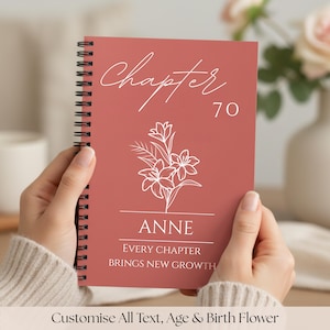 May include: A coral-coloured spiral-bound notebook with the word "Chapter" in script, the number 70, and the name "ANNE". A white floral illustration and the text "EVERY CHAPTER BRINGS NEW GROWTH" are displayed. The notebook is held by a person.