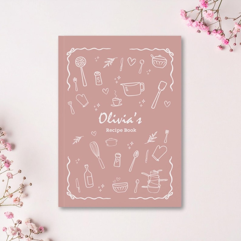 Puede incluir: Un libro de recetas rosa empolvado con el t&iacute;tulo "Olivia's Recipe Book" en letra cursiva. La portada est&aacute; decorada con dibujos lineales blancos de utensilios de cocina y alimentos. Flores rosas en el fondo.