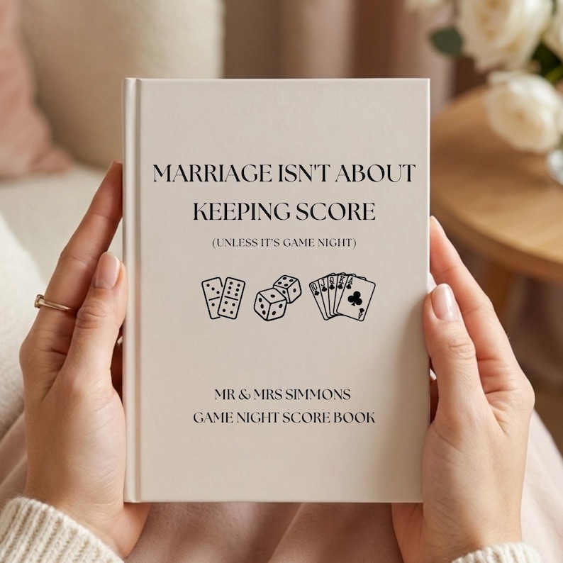 May include: A cream-colored hardcover score book with black text that reads: MARRIAGE ISN'T ABOUT KEEPING SCORE (UNLESS IT'S GAME NIGHT), MR & MRS SIMMONS, GAME NIGHT SCORE BOOK, featuring black line art icons of dominoes, dice, and playing cards.