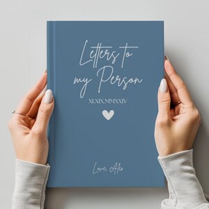 以下が含まれることがあります： 白いスクリプトで「Letters to my Person」というタイトルの青いジャーナル。表紙には白いハートと日付XI.XIX.MMXXIVが含まれています。ジャーナルはマニキュアされた爪を持つ2つの手で保持され、署名「Love, Alex」が一番下にあります。