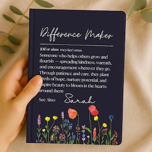 Puede incluir: Un diario azul marino con las palabras "Difference Maker" en escritura blanca. La definición de "Difference Maker" está impresa debajo del título. La parte inferior del diario presenta un diseño floral colorido.