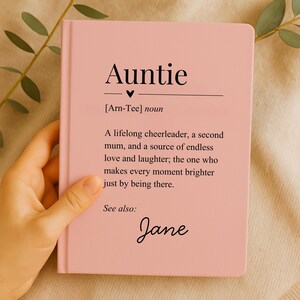 Peut inclure: Un carnet rose avec le mot "Auntie" en haut, suivi d'une définition. La définition décrit une tante comme une pom-pom girl à vie. Le nom "Jane" est écrit en bas.