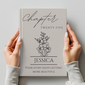 May include: A personalized journal with a light gray cover held by two hands. The cover features the word "Chapter" in cursive, "Twenty Five", a floral illustration, the name "Jessica", and the quote "Your story keeps getting more beautiful."