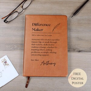 May include: A brown leather journal with the words "Difference Maker" embossed on the cover, along with a definition and the name "Anthony." A feather illustration and a ribbon bookmark are also visible. The journal is on a wooden surface.