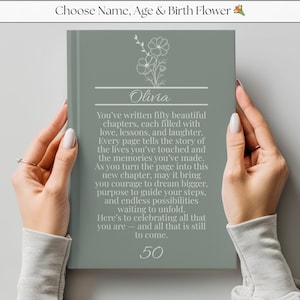 May include: A sage green journal held by two hands, featuring the name "Olivia" in script, floral illustrations, and the number "50". The cover includes text celebrating life's chapters. The journal is a thoughtful gift.