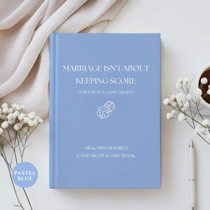 May include: A pastel blue hardcover book with white text that reads MARRIAGE ISN'T ABOUT KEEPING SCORE (UNLESS IT'S GAME NIGHT) and MR & MRS MOORE'S GAME NIGHT SCORE BOOK, featuring a simple white line drawing of two dice in the center.