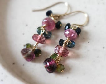 May include: A pair of gold-toned earrings with clusters of colorful gemstones. The earrings feature pink, green, and blue tourmaline beads, suspended from delicate gold hooks. The earrings are displayed on a white, speckled surface.