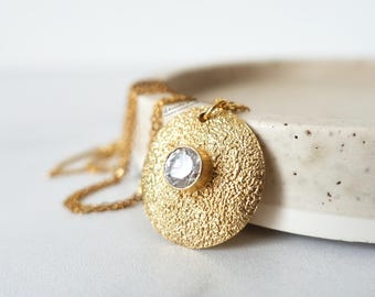 May include: A gold-colored necklace with a textured, circular pendant. The pendant features a central, clear gemstone. The necklace is displayed on a white surface, showcasing the jewelry's design and craftsmanship.
