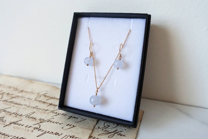 May include: A delicate gold necklace and matching earrings set with light blue gemstone pendants. The jewellery is displayed in a black box with white lining.
