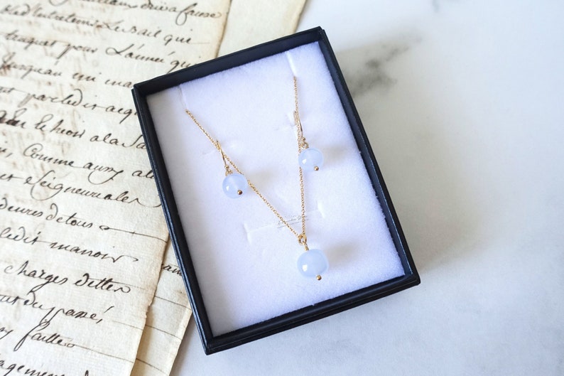May include: A delicate gold necklace and matching earrings with light blue round gemstone pendants. The jewellery is displayed in a black velvet box.