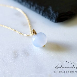 May include: A delicate gold chain necklace with a light blue gemstone pendant. The pendant is a smooth, rounded shape and is set in a simple gold wire wrap.