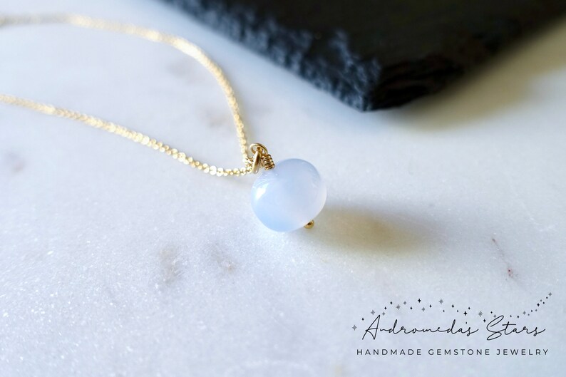 May include: A delicate gold chain necklace with a light blue gemstone pendant. The pendant is a smooth, rounded shape and is set in a simple gold wire wrap.