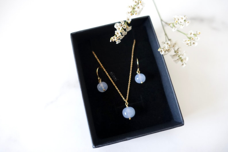 May include: A set of delicate jewellery featuring a light blue gemstone pendant necklace and matching earrings. The necklace and earrings have a gold chain and are displayed in a black box.