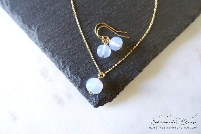 May include: A delicate gold chain necklace and matching earrings with a light blue round gemstone pendant. The jewellery is displayed on a dark grey surface.