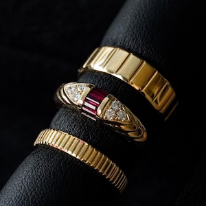 Puede incluir: Tres anillos de oro apilados sobre una superficie negra. El anillo superior presenta una piedra preciosa roja rodeada de pequeños diamantes. El anillo del medio es una banda lisa con una superficie texturizada. El anillo inferior es una banda lisa con una superficie texturizada.