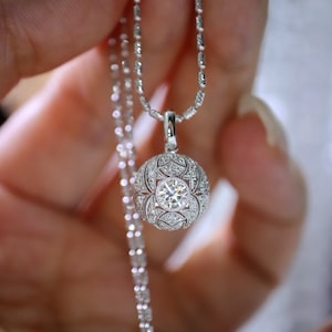 Può includere: Una collana d'argento con un ciondolo rotondo che presenta un grande diamante trasparente circondato da diamanti più piccoli in un delicato motivo floreale.