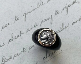Puede incluir: Un anillo de ónix negro con una banda dorada y un camafeo de plata con el perfil de una mujer. El camafeo está engastado en un marco circular.