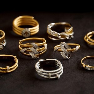 Pu&ograve; includere: Una collezione di nove anelli in oro e argento con disegni intricati e diamanti scintillanti. Gli anelli sono disposti su una superficie nera.