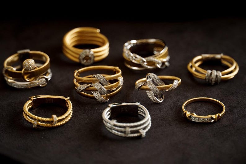 Pu&ograve; includere: Una collezione di nove anelli in oro e argento con disegni intricati e diamanti scintillanti. Gli anelli sono disposti su una superficie nera.