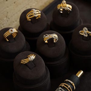 Pu&ograve; includere: Una collezione di anelli in oro con accenti di diamanti esposti su porta anelli in velluto nero. Gli anelli presentano vari design, tra cui un cerchio, una catena e un nodo.