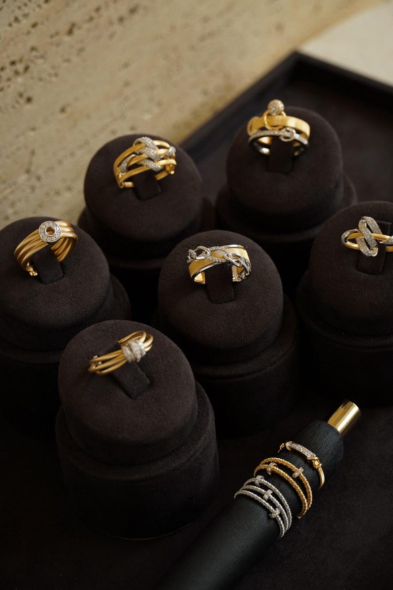 Pu&ograve; includere: Una collezione di anelli in oro con accenti di diamanti esposti su porta anelli in velluto nero. Gli anelli presentano vari design, tra cui un cerchio, una catena e un nodo.