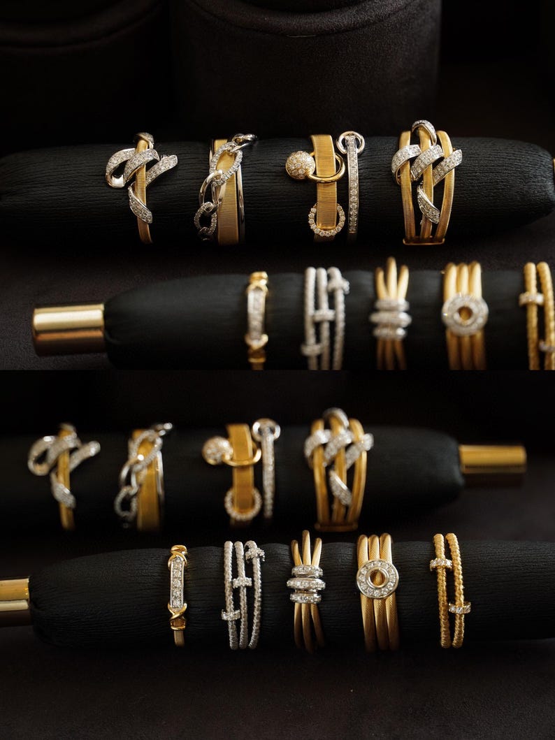 Pu&ograve; includere: Una collezione di anelli in oro e argento con dettagli in diamanti, esposti su velluto nero. Gli anelli presentano diversi design, tra cui un anello a catena, un nodo e un cerchio.