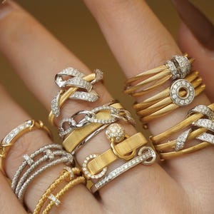 Pu&ograve; includere: Una mano che indossa una pila di anelli in oro e argento con accenti di diamanti. Gli anelli presentano vari design, tra cui un anello a catena, una corda ritorta e un anello con una piccola palla in cima.