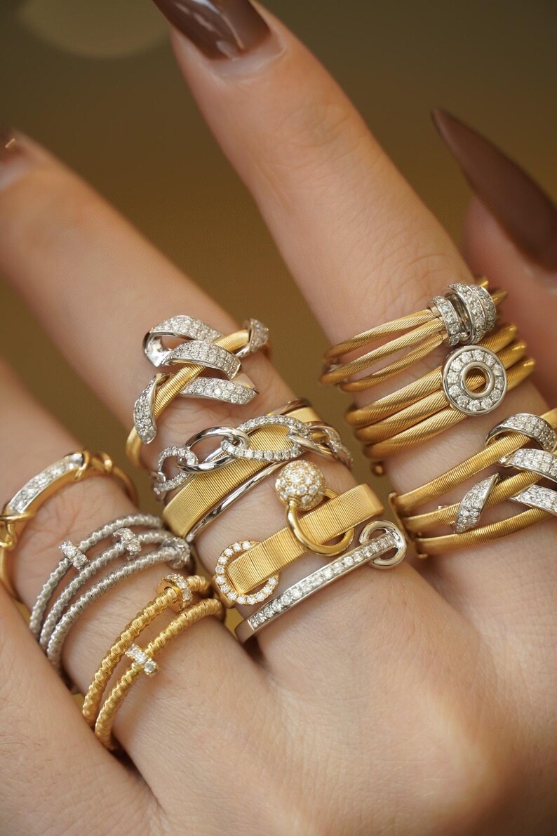 Pu&ograve; includere: Una mano che indossa una pila di anelli in oro e argento con accenti di diamanti. Gli anelli presentano vari design, tra cui un anello a catena, una corda ritorta e un anello con una piccola palla in cima.