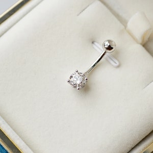Può includere: Un anello per l'ombelico in argento con una pietra rotonda trasparente in una scatola di velluto bianco.