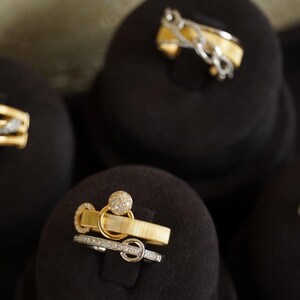 Pu&ograve; includere: Quattro anelli d'oro con accenti di diamanti esposti su cuscini di velluto nero. Gli anelli sono di stili diversi, tra cui una fascia attorcigliata, un anello con una pietra rotonda e un anello con un design a catena delicato.