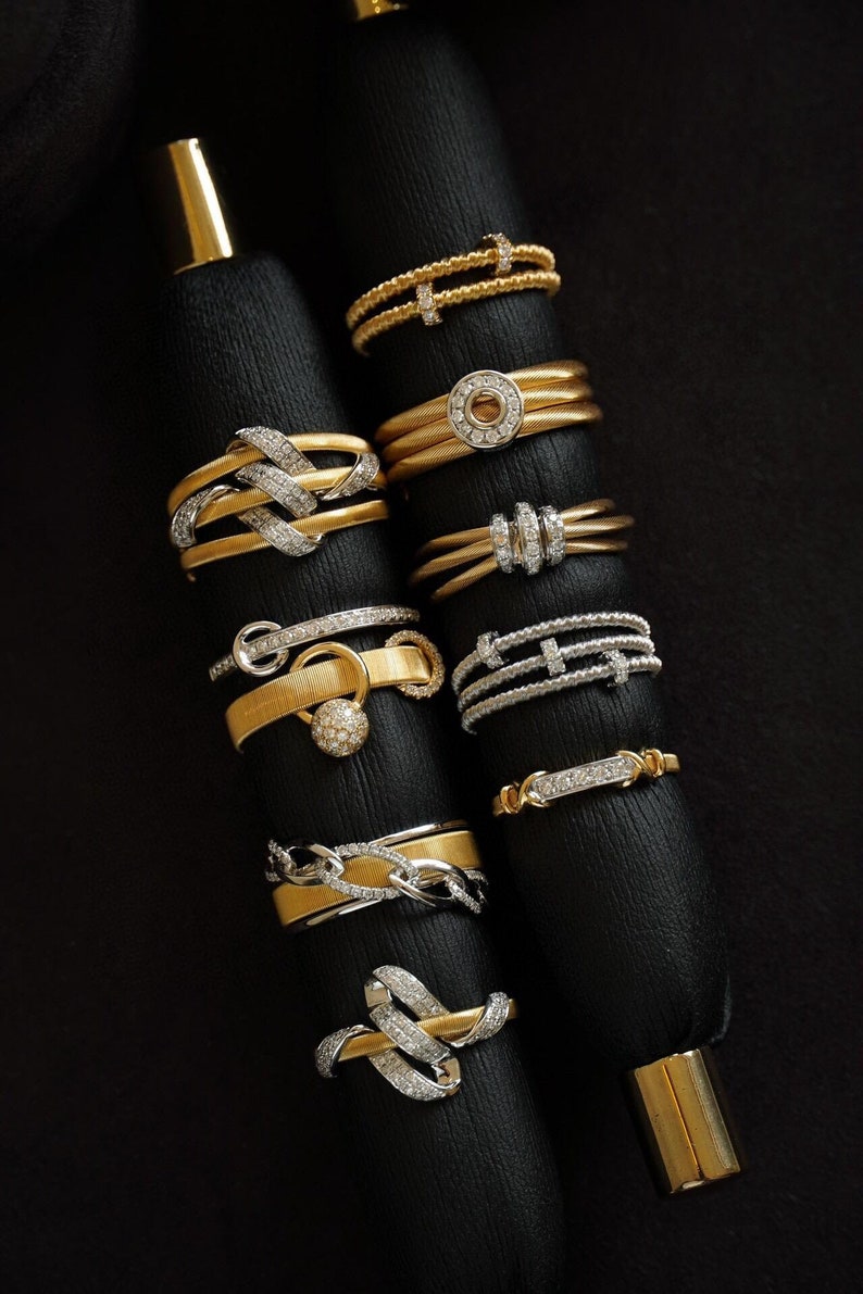 Pu&ograve; includere: Una collezione di anelli in oro e argento con dettagli in diamanti, esposti su supporti in velluto nero. Gli anelli presentano diversi design, tra cui un cerchio con un diamante al centro, un design intrecciato e un anello con una piccola sfera di diamanti.