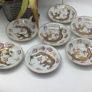 Op de afbeelding: Set van zes kleine witte porseleinen schoteltjes met gouden en rode drakenontwerpen.