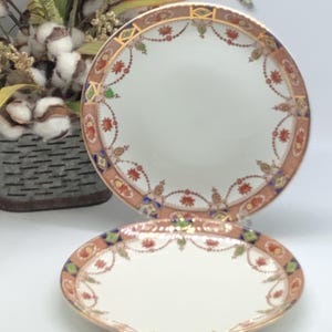 May include: Two vintage porcelain plates with a decorative floral pattern in shades of red, blue, green, and gold. The plates have a white background and a gold rim.