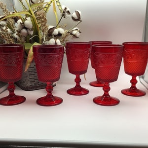 May include: Six red glass goblets with a diamond pattern and floral design. The goblets are arranged in a row on a white surface.