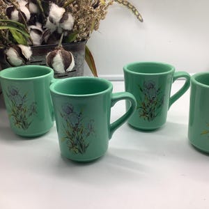 Peut inclure: Quatre tasses en céramique vertes avec un motif floral d'iris. Les tasses ont une poignée incurvée et une finition lisse et brillante. Le motif d'iris présente des fleurs violettes et jaunes avec des tiges et des feuilles vertes.