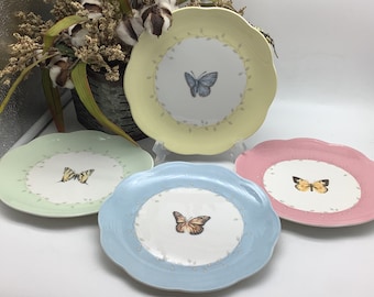 May include: Set of four decorative plates with scalloped edges. Each plate features a different colored rim and a butterfly design in the center. The plates are yellow, green, blue, and pink.