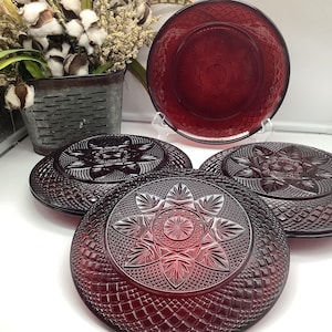 May include: A set of four round, deep red glass plates featuring a pressed pattern with a central starburst motif surrounded by a textured diamond grid design. The plates are translucent, showing light through the glass, and have a raised rim.