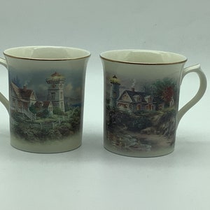 Può includere: Due tazze in ceramica bianca con bordo dorato. Ogni tazza presenta un dipinto diverso di un faro e una casa con uno sfondo panoramico.
