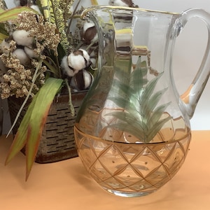 May include: A clear glass pitcher with a gold rim and a pineapple design. The pitcher has a handle and is decorated with green leaves and a gold diamond pattern. A decorative basket with cotton and greenery is in the background.
