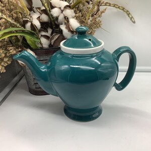 Può includere: Teiera in ceramica color verde acqua con bordo e coperchio bianchi. La teiera ha un corpo arrotondato, un manico curvo e un beccuccio. Il coperchio ha un piccolo pomello. La teiera è su una superficie bianca.