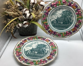 May include: Two vintage ceramic plates with a green and white design featuring a cottage scene. The plates have a scalloped edge and are decorated with a colorful floral and fruit border.