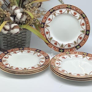 May include: A set of fine china plates with a white center and a decorative border in shades of orange, navy, green, and gold. The plates are stacked and arranged, showcasing their intricate design. A floral arrangement is in the background.