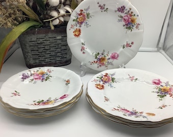 May include: A collection of white porcelain plates with a scalloped edge and gold trim. Each plate features a floral design in pink, yellow, orange, and purple. The plates are stacked in sets of three.