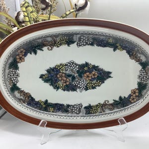 May include: An oval ceramic serving platter with a wide, solid brown rim. The center features a white background decorated with a central cluster and a surrounding border of colorful grapes, green leaves, and small flowers in shades of blue and yellow.